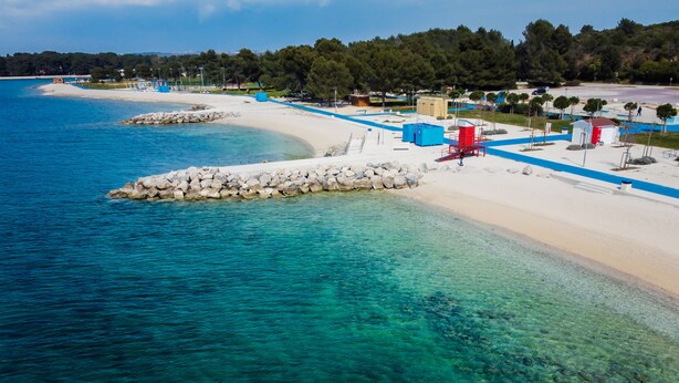 Una playa costera con aguas turquesas cristalinas, una orilla arenosa y un pequeño edificio con techo rojo.