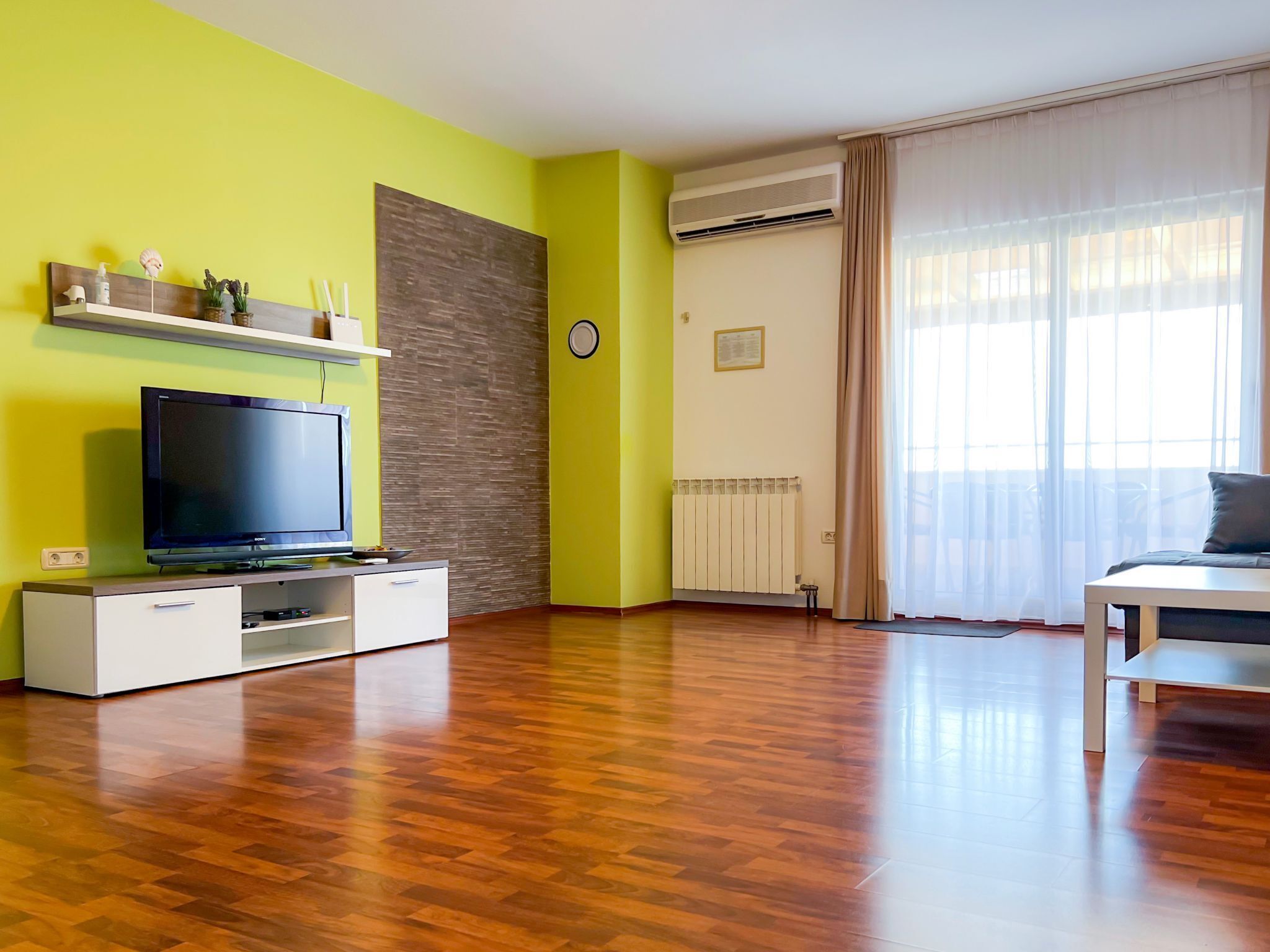 Ein modernes Wohnzimmer mit Flachbildfernseher, Holzboden, Wandgerätet Klimaanlage und einem großen Fenster mit durchsichtigen Vorhängen.