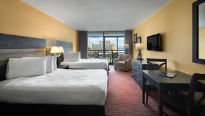 Ein Hotelzimmer mit zwei Betten, einem Schreibtisch, einem Stuhl, einem Fernseher und einem Balkon mit Blick auf die Stadt.