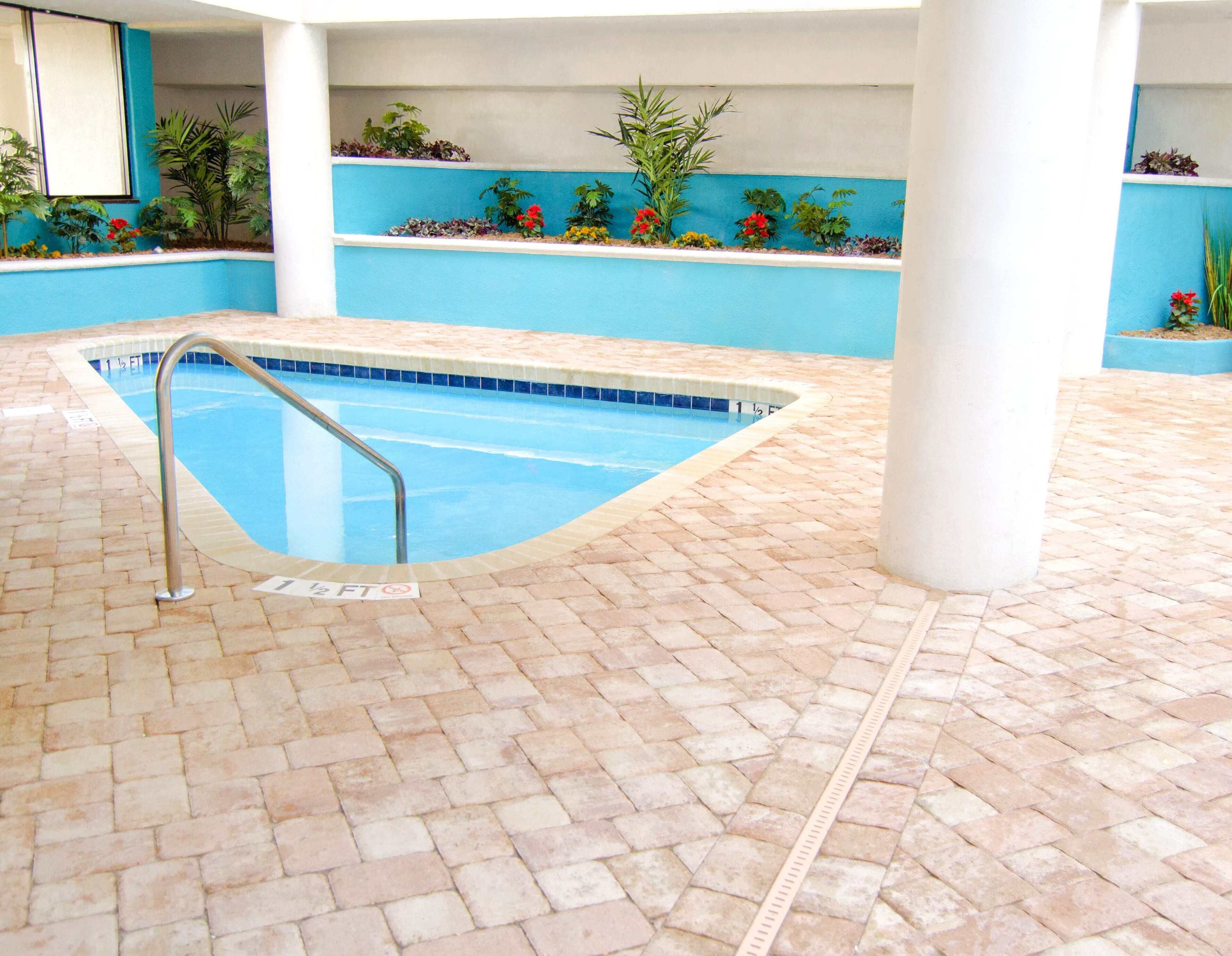 An indoor swimming pool with a tiled floor, a metal pool ladder, and plants along the walls.
