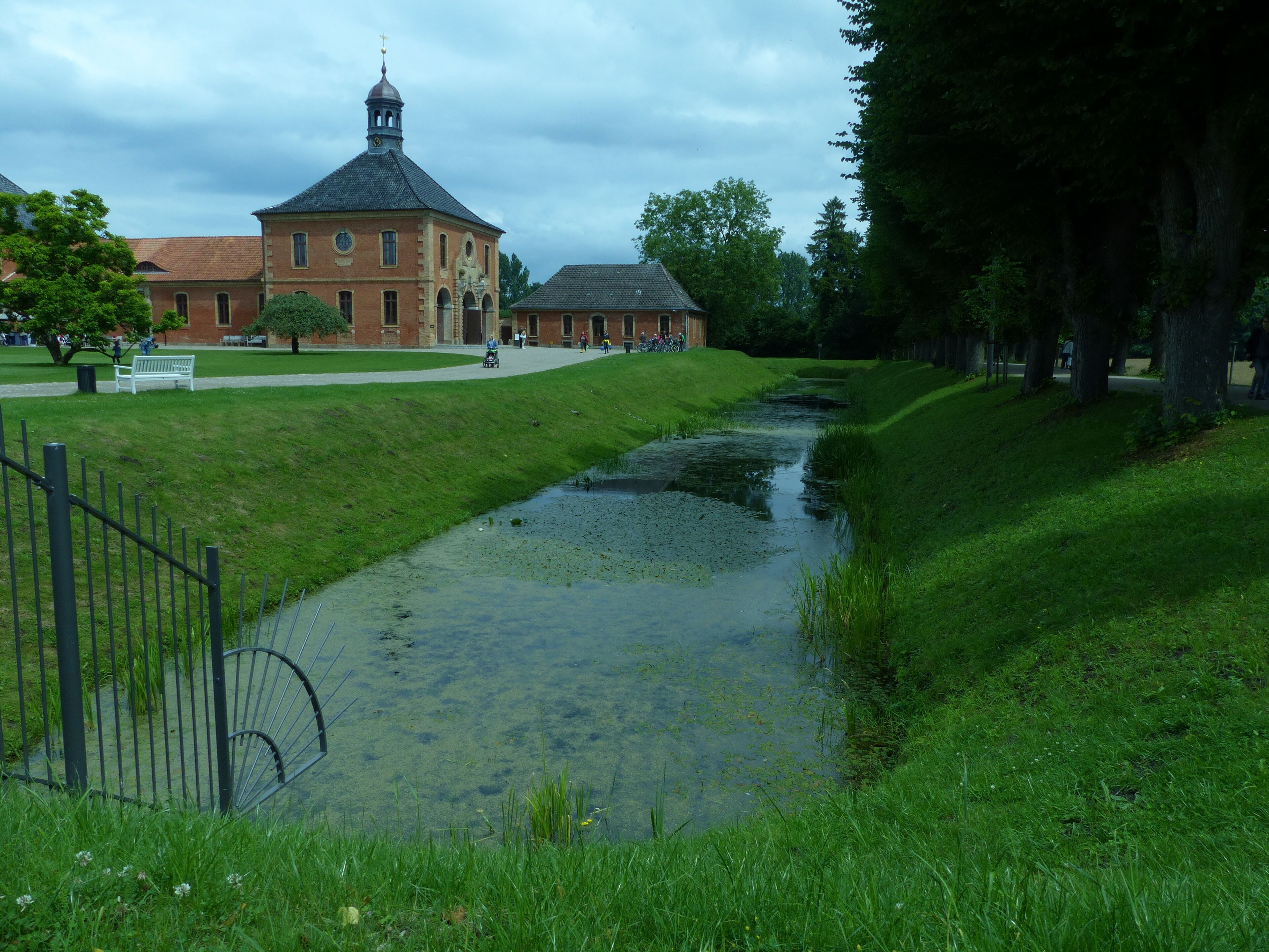 A grassy pathway with a small water channel, a brick building with a tower, and a bench in the foreground.