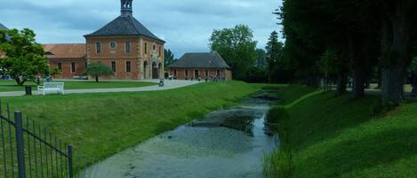 A grassy pathway with a small water channel, a brick building with a tower, and a bench in the foreground.