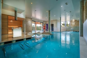An indoor swimming pool with clear water, a reception area with a white counter, and a view of the outside through large windows.