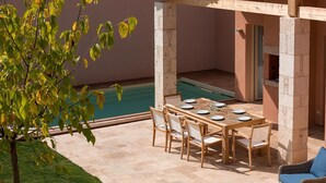 A covered patio with a pool, wooden table and chairs, and a blue cushioned chair.