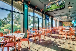 Un restaurante espacioso con amplias ventanas, sillas anaranjadas y una pantalla de televisión mostrando un partido deportivo.