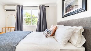 Un lit bien fait, avec du linge de lit blanc et une couverture grise, une tête de lit grise et un tableau encadré accroché au mur.