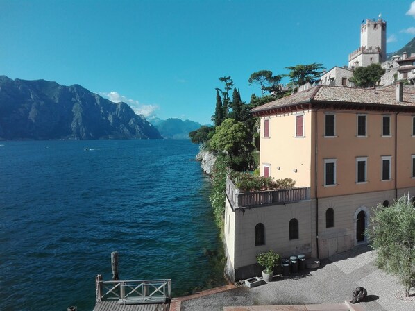 A coastal building with a balcony overlooking a lake, a tower, and mountains in the background.