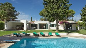 Ein Poolbereich mit einem klaren blauen Pool, grünen Liegestühlen und einem weißen Gebäude mit einer überdachten Terrasse.