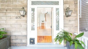 A brick house entrance with a number '405' displayed on the wall, a wooden staircase leading inside, and two wall-mounted lanterns.