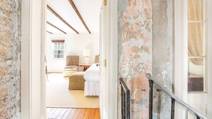 A room with exposed brick walls, a wooden ceiling, a bed with white bedding, a lamp, and a window with curtains.
