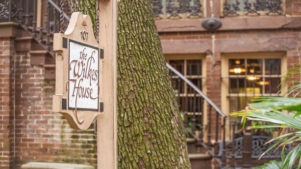 A wooden sign with "the Wilkes House" written on it, attached to a tree in front of a brick building with windows and a staircase.