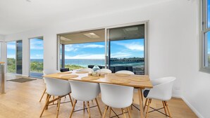 A dining area with a wooden table, white chairs, and large windows offering a view of the sea and a bridge. - Seascape, Bombo, South Coast (Bombo)