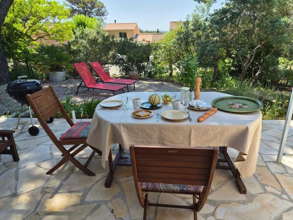 An outdoor dining area with a round table set for four, surrounded by stone flooring, wooden chairs, and red lounge chairs.