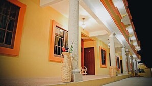 Un hotel con fachada amarilla, múltiples ventanas con marcos anaranjados y un espacio exterior cubierto con columnas y cestas de mimbre.