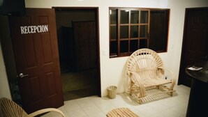 Zona de recepción con puerta de madera que dice 'RECEPCIÓN', muebles de mimbre y una ventana con persianas.