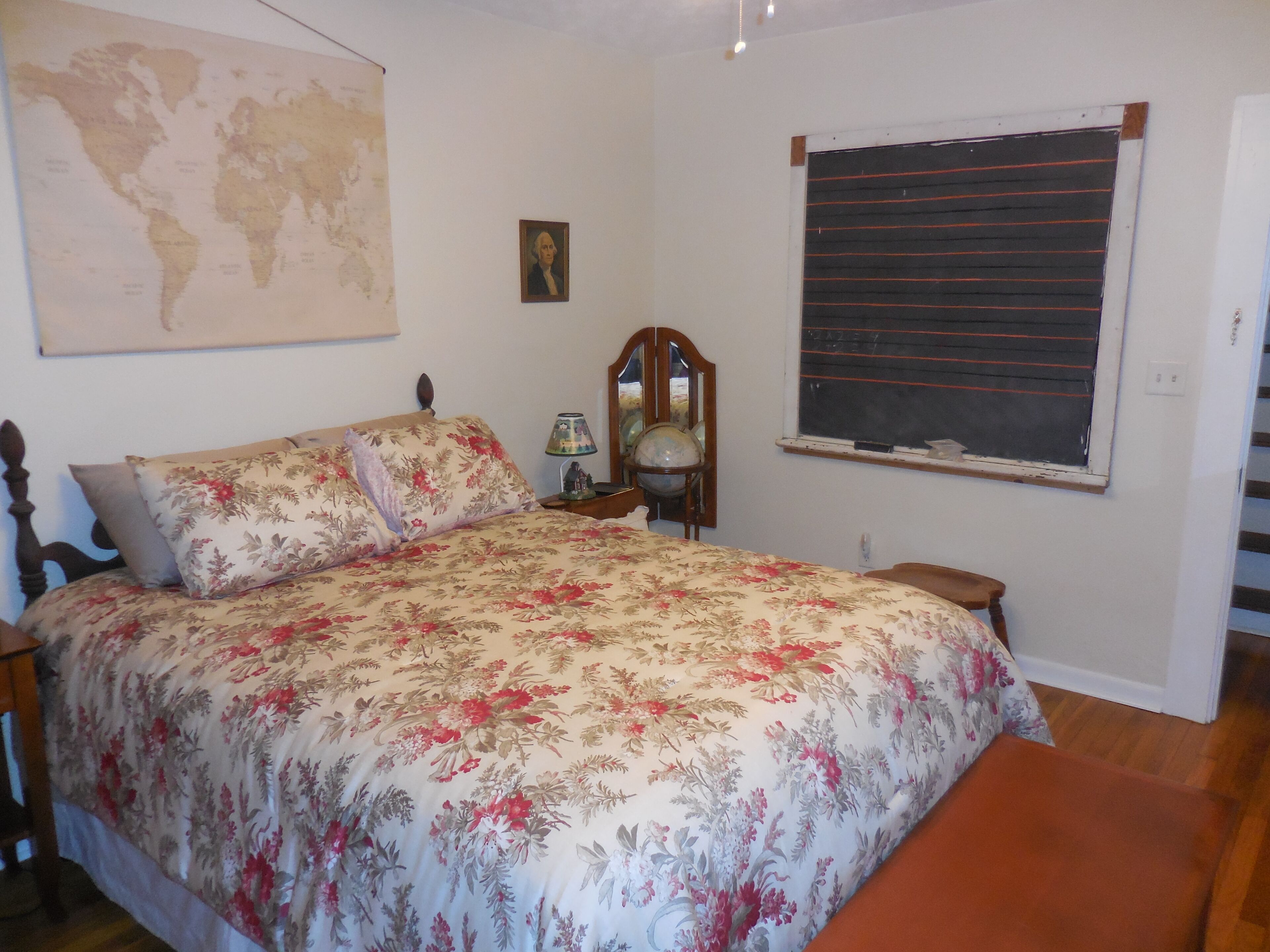 A bedroom with a bed, a world map, a framed picture, a globe, a window with blinds, and a wooden bench.