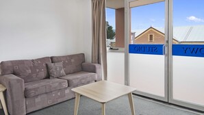 Una habitación con un sofá gris estampado, una pequeña mesa de madera y un ventanal corredero que da a un espacio al aire libre.