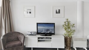 Una sala moderna con un televisor de pantalla plana sobre un mueble blanco, un sillón gris y una planta en maceta.
