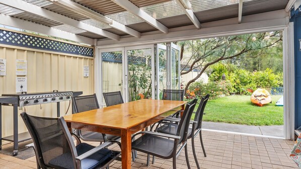 An outdoor patio with a wooden table and chairs, a barbecue grill, and a view of a garden.