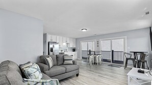 Un salon moderne avec un canapé gris, un coin repas avec une table et des chaises blanches, et une cuisine en arrière-plan.