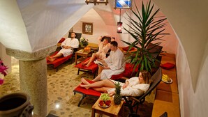 Un cuarto de spa con cuatro personas relajándose en sillones rojos, una mesa con frutas y una taza, y una planta grande.