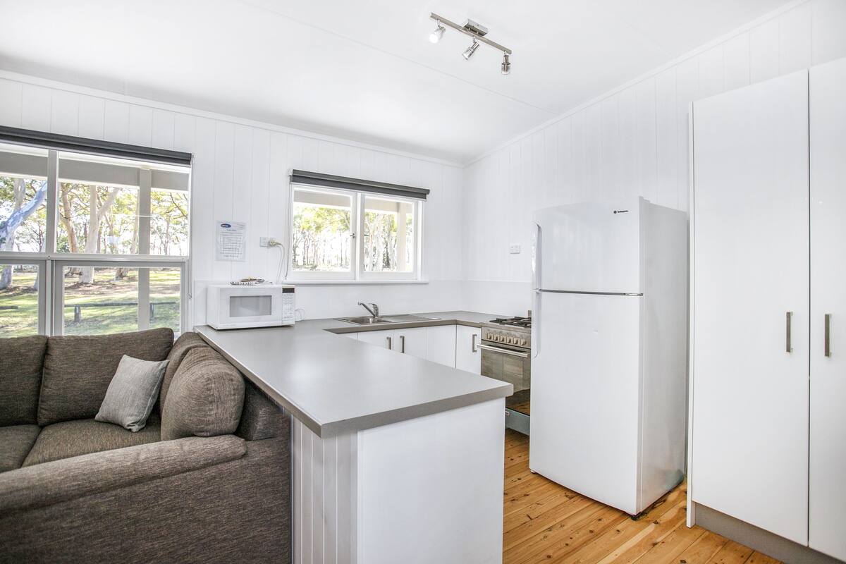 Eine moderne Küche mit weißen Schränken, einer eingebauten Mikrowelle und einem Kühlschrank. Ein Holzboden und ein Fenster sorgen für natürliches Licht.