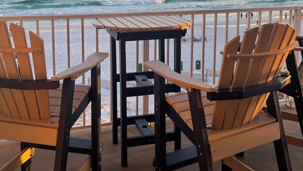Un balcon avec des chaises et une table en bois, d’où l’on peut admirer l’océan au coucher du soleil.