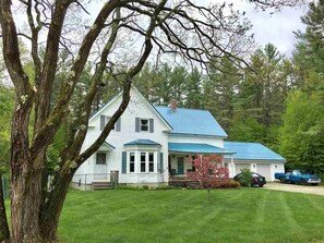 Une maison blanche aux bordures et au toit bleus, entourée d’une pelouse bien entretenue et d’arbres.