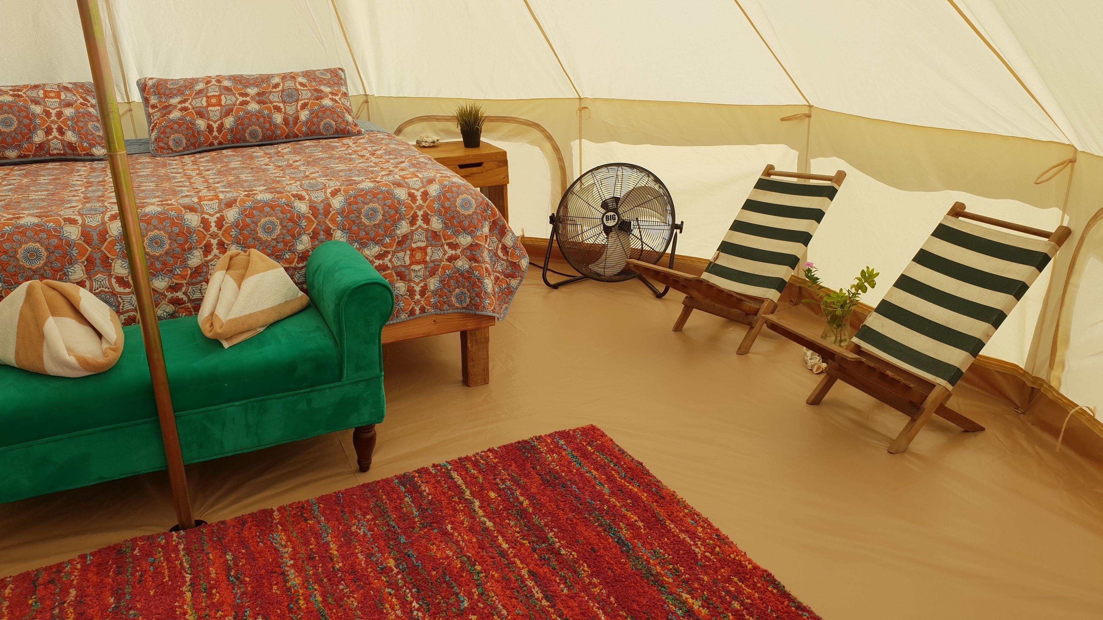 Ein Zeltraum mit einem Bett, einem grünen Sessel, gestreiften Liegestühlen und einem Ventilator.