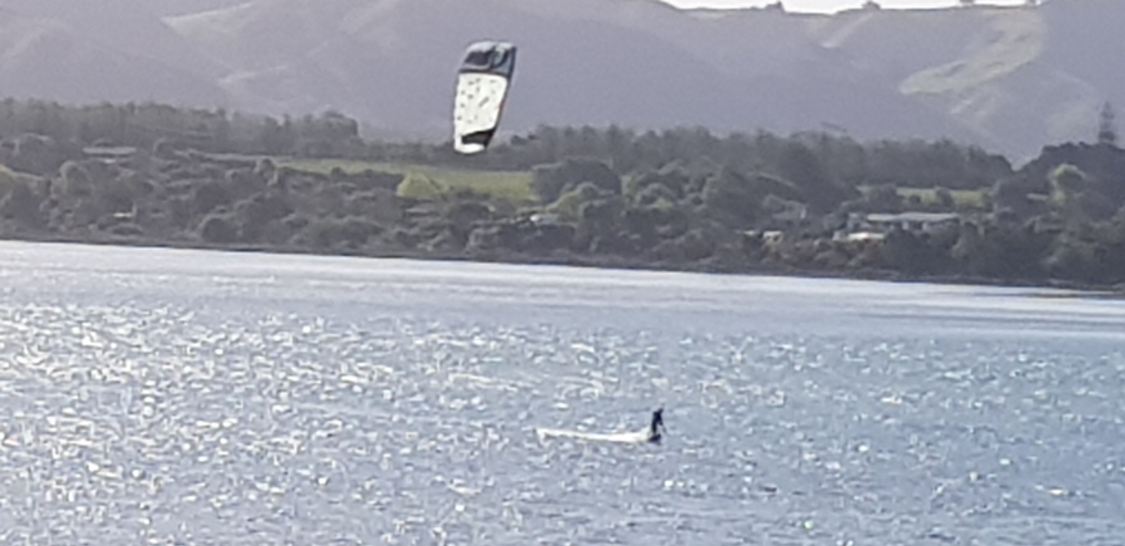 Una persona practica kitesurf en un cuerpo de agua, con un paisaje montañoso al fondo.