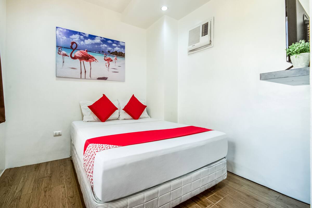 Une chambre à coucher avec un lit, un tableau représentant des flamants roses, un coussin rouge et une petite plante posée sur une étagère.