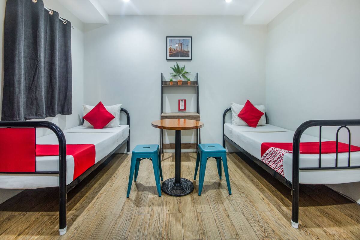 Une chambre avec deux lits simples, une petite table, deux tabourets, une étagère avec des plantes et un tableau encadré au mur.
