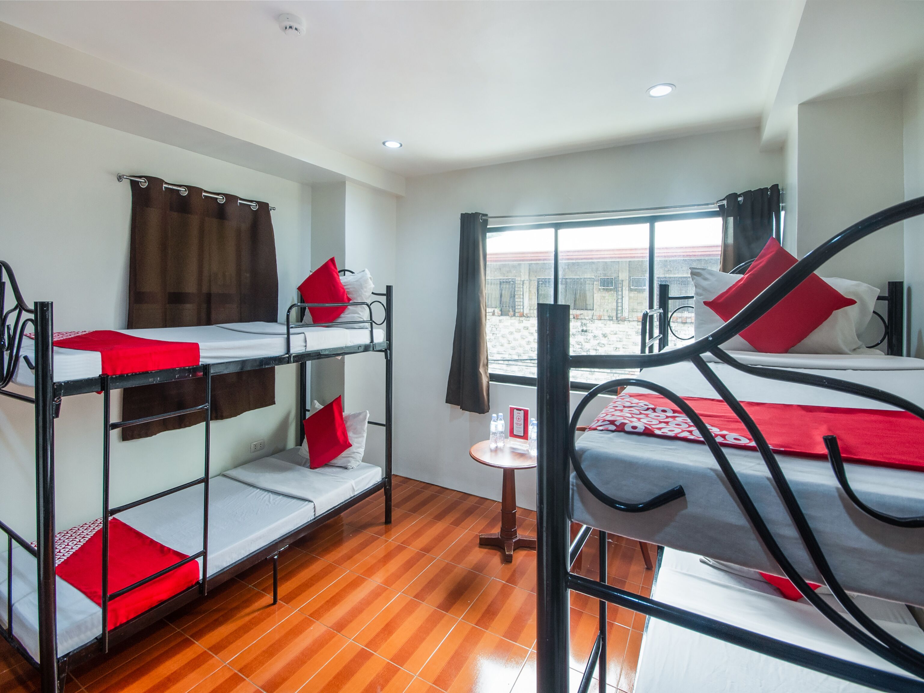 A dormitory room with bunk beds, a small table with a chair, and a window with a view of the outside.