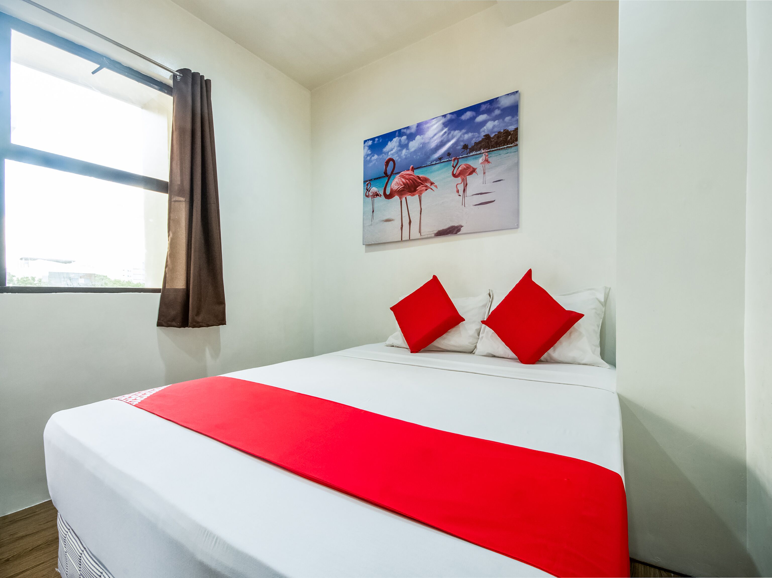 Habitación de hotel con cama, almohadas rojas, un cuadro de flamencos y una ventana con cortinas.