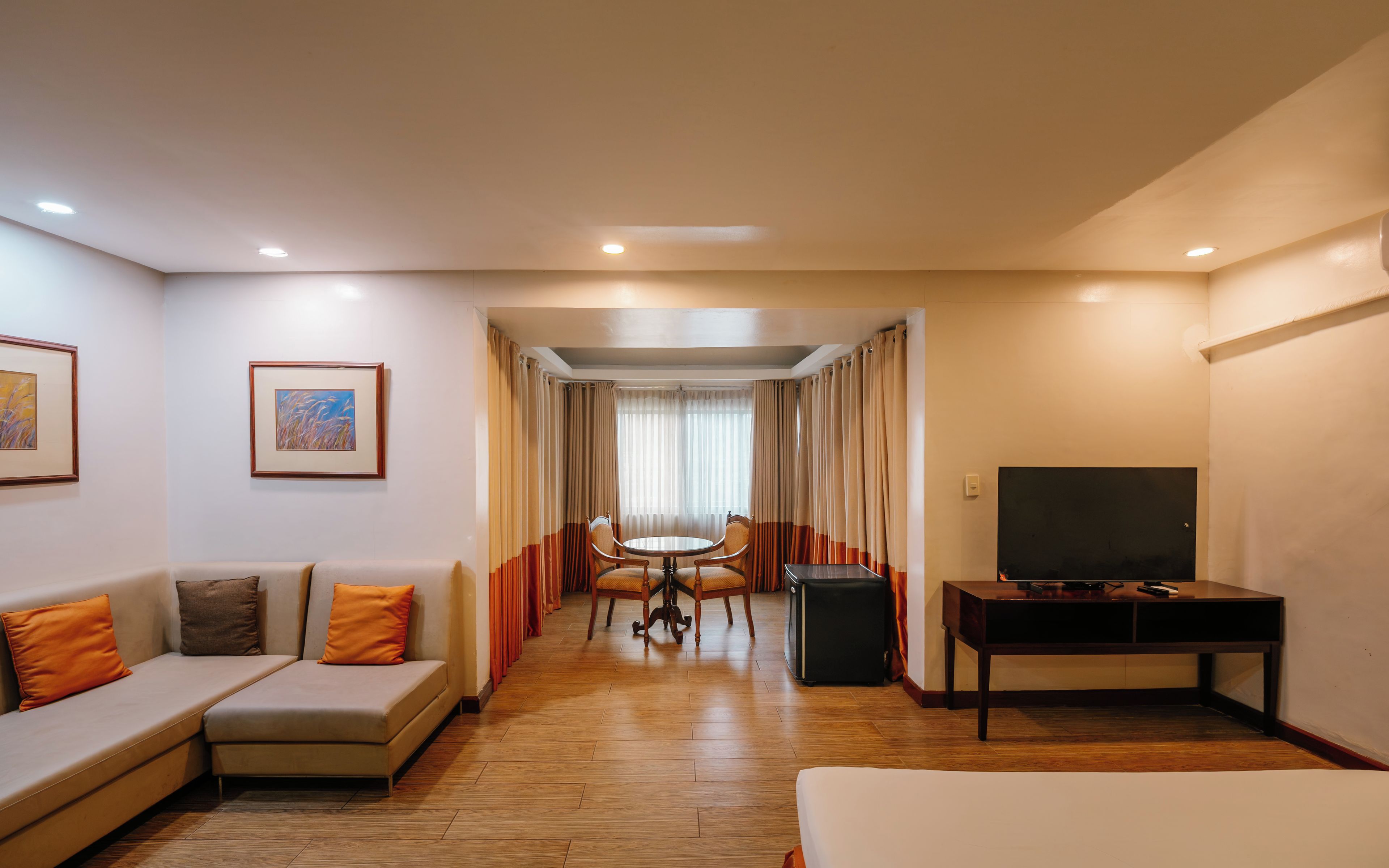 A hotel room with a sofa, a TV, a dining table with chairs, and a window with curtains.