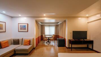 A hotel room with a sofa, a TV, a dining table with chairs, and a window with curtains.