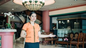 A woman in a uniform standing in a lobby with a reception desk, chairs, and a staircase in the background. - La Fiesta Hotel (Iloilo)
