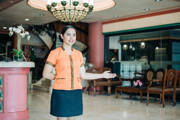 A woman in a uniform standing in a lobby with a reception desk, chairs, and a staircase in the background. - La Fiesta Hotel (Iloilo)