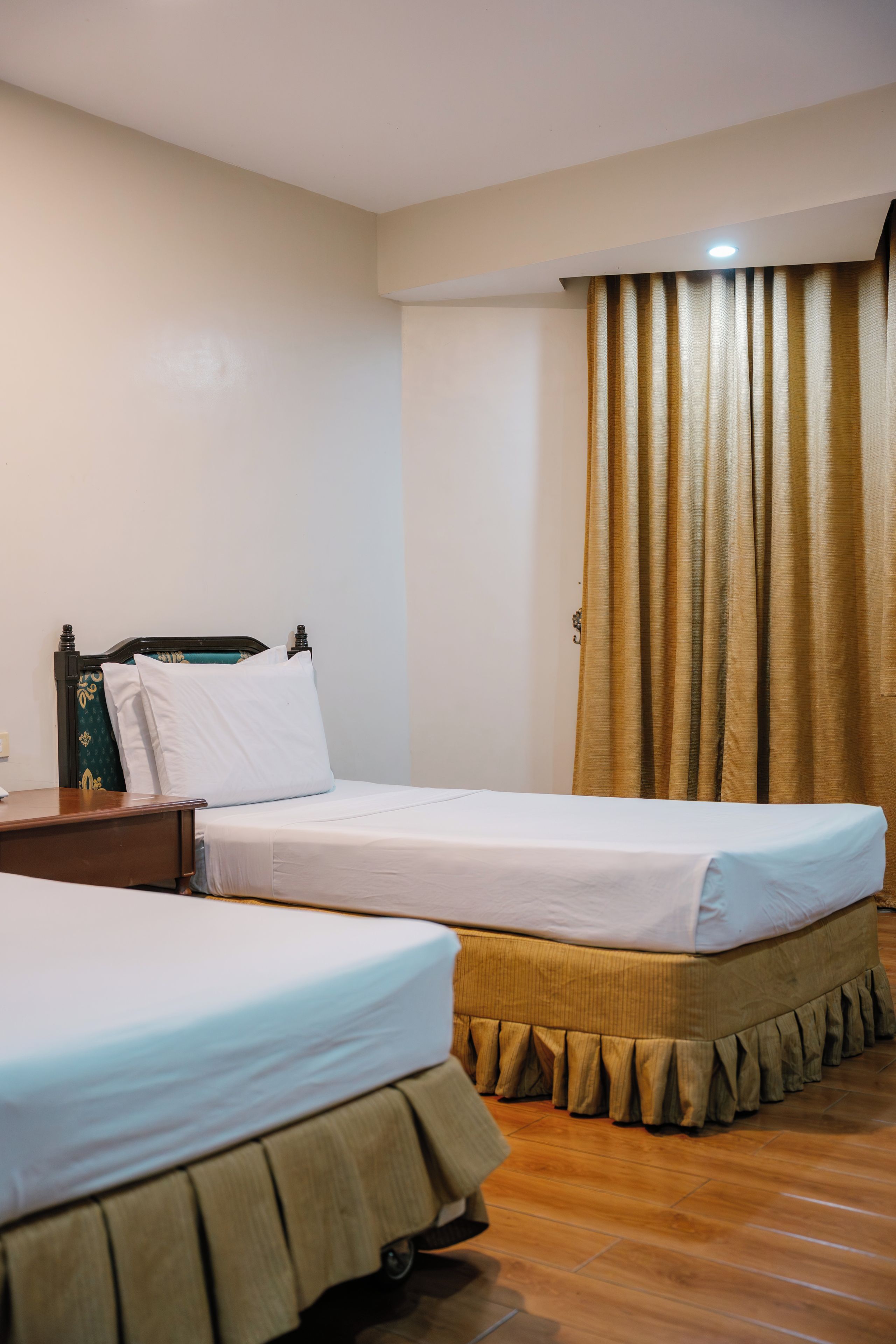 A hotel room with two single beds, a wooden nightstand, a small table, and a curtain.