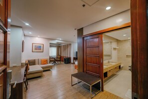 A hotel room with a wooden floor, a sofa, a small table, a bed with a wooden headboard, and a bathroom with a large mirror. - La Fiesta Hotel (Iloilo)