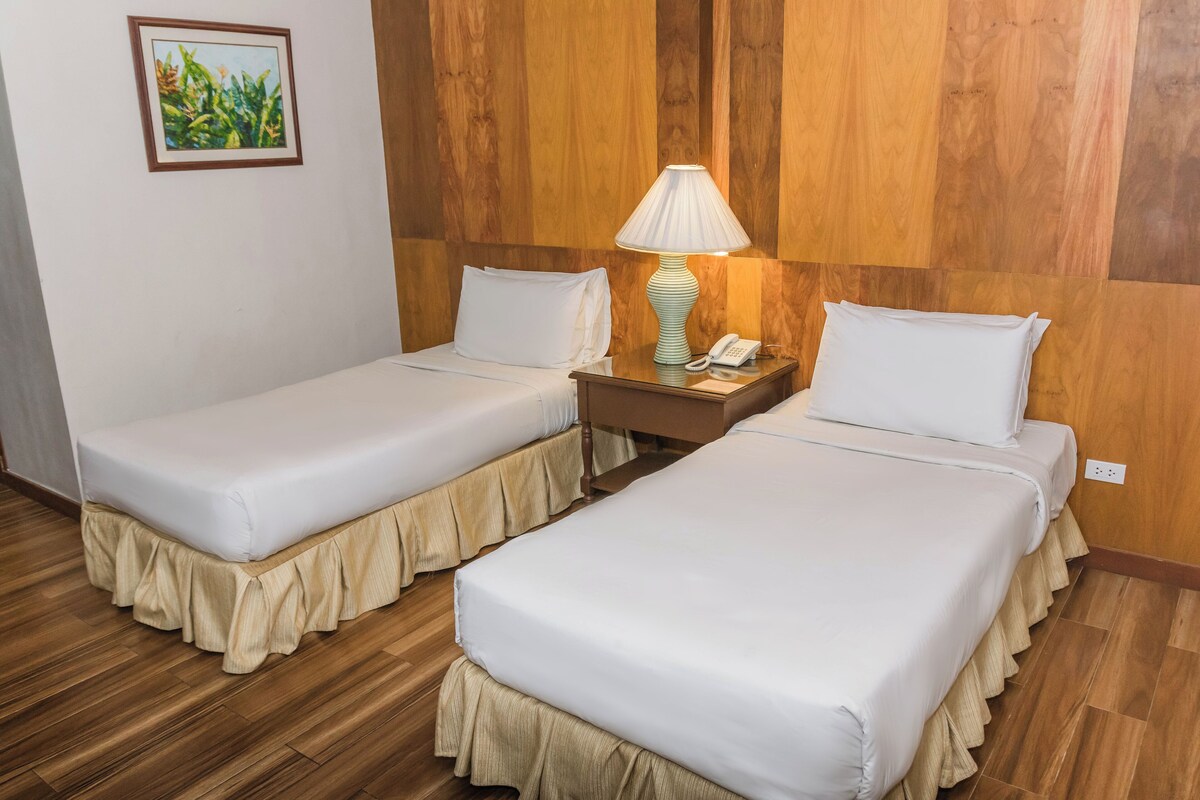 Une chambre d’hôtel avec deux lits simples, une table de chevet avec une lampe et un téléphone, des murs recouverts de panneaux de bois et un tableau encadré accroché au mur.