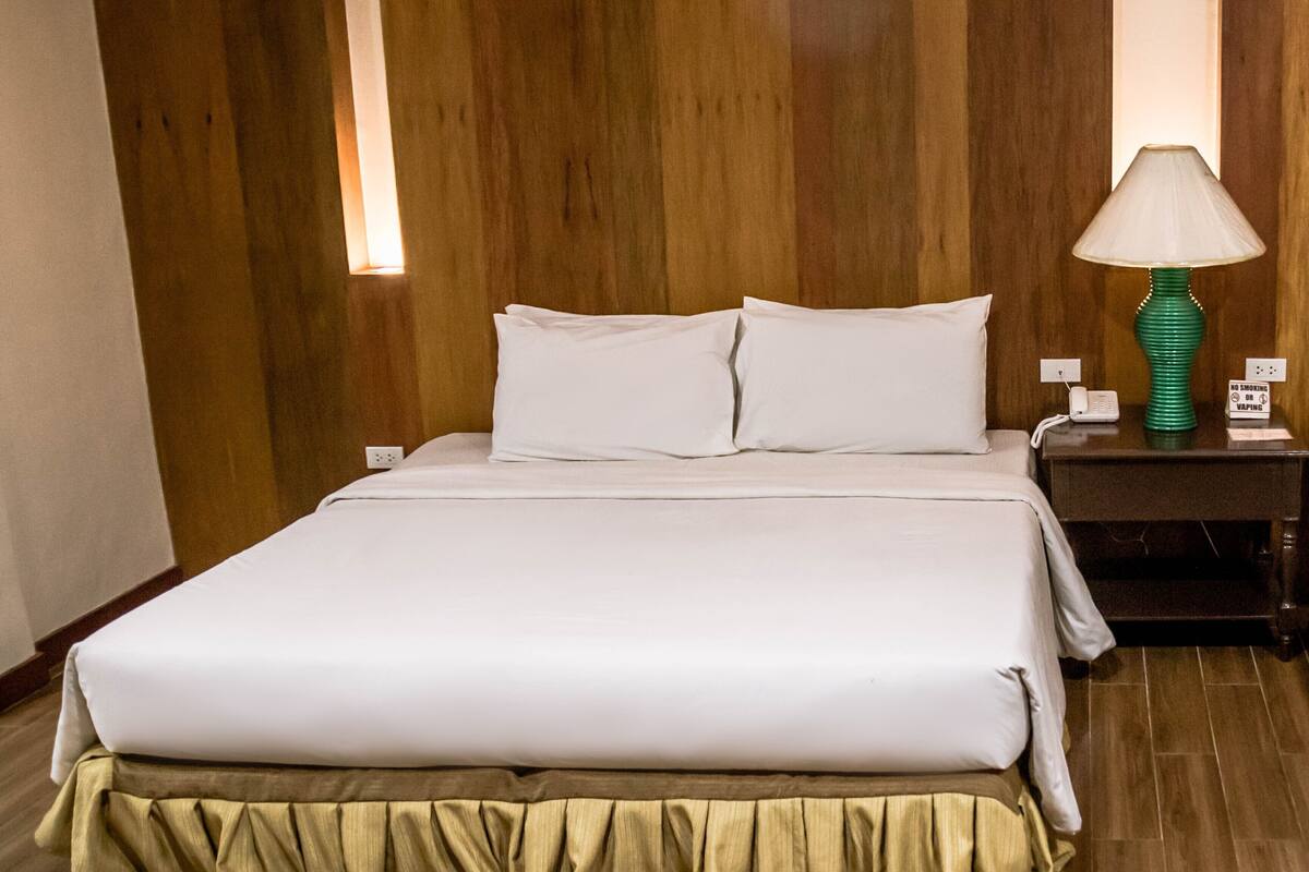 A neatly made bed with white linens, a green lamp on a bedside table, and a wooden floor.