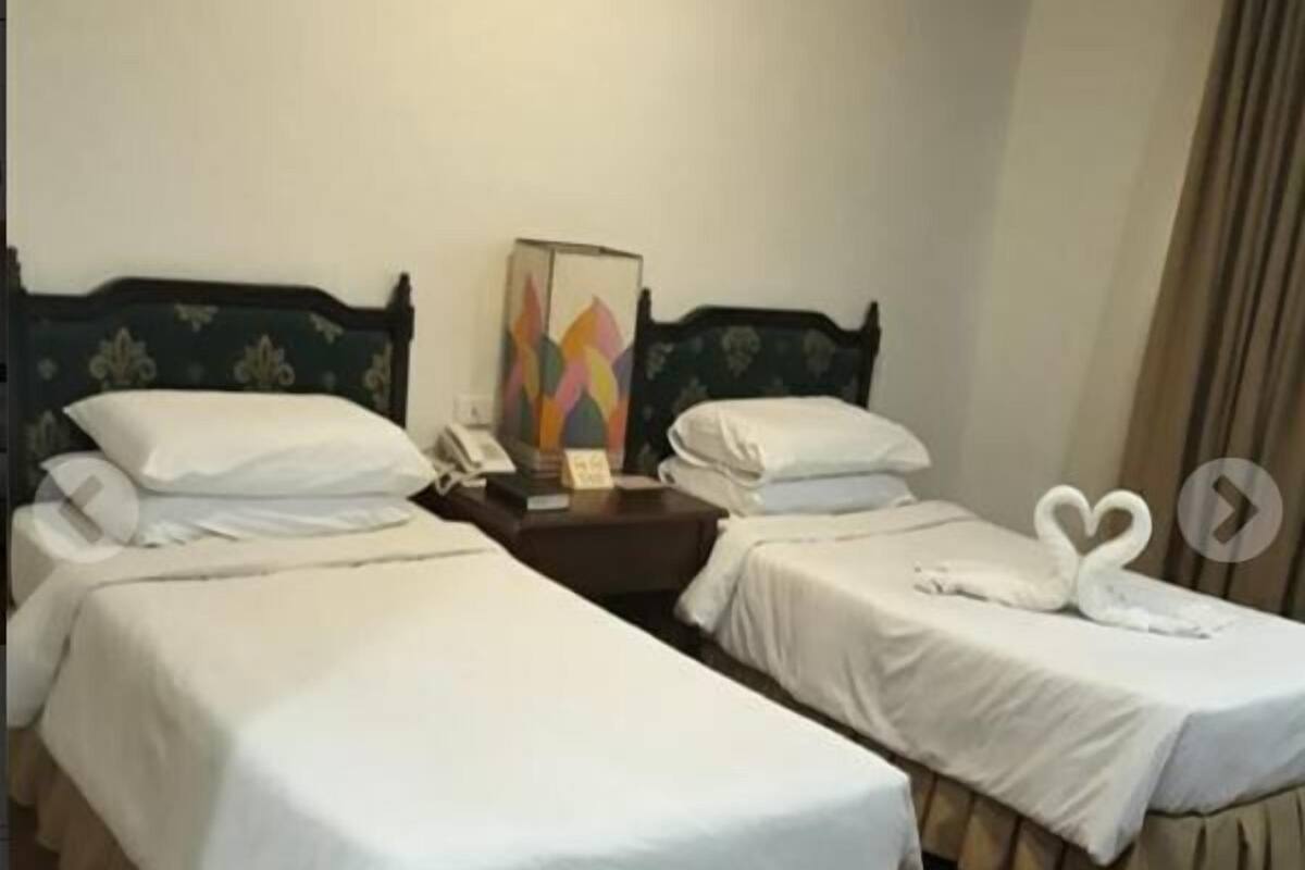 A hotel room with two single beds, a bedside table with a lamp, a small table with a vase, and a painting on the wall.
