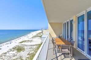 En balkong med ett träbord och stolar, med utsikt över en strand med parasoller och folk.