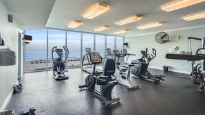 Un gimnasio con equipo de cardio, un ventilador en la pared y vista a la playa.