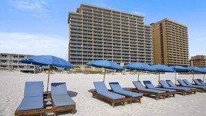 Una playa con hileras de tumbonas de madera y sombrillas azules, un gran edificio de hotel al fondo.