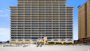 Un hotel de varios pisos con una fachada uniforme, múltiples balcones y una playa arenosa al frente.
