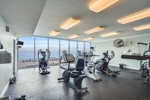 Een sportschool met cardioapparatuur, een ventilator aan de muur en uitzicht op het strand.