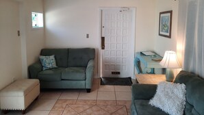 A living room with a green sofa, a smaller green armchair, a wooden side table with a lamp, and a door with a window.