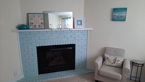 A living room with a blue brick fireplace, a beige armchair with a bird pillow, and a small round table.